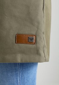 Olivově zelená plátěná tkanina s hnědým koženým pásem s kovovým logem. Modrý denim viditelný pod lemem oděvu.