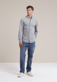 Camisa de botones de manga larga gris con collar y detalle de bolsillo, combinada con jeans azules y zapatillas blancas, de pie contra un fondo neutro.