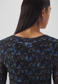 Femme aux longs cheveux noirs portant un haut bleu et noir à motifs floraux avec "BOSS" brodé sur le haut du dos, montrant son oreille droite ornée de boucles d'oreilles créoles.