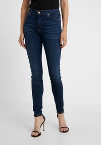Jeans skinny azul oscuro de denim, con una cintura alta, cinco bolsillos y una cremallera con botón. Combinados con tacones negros de tiras.