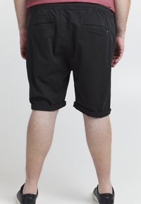 Homme portant un short noir retroussé et des baskets noires, debout de dos sur un fond uni.