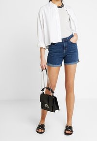 Džínové šortky světle modré barvy s roztřepeným lemováním. Bílá košile na knoflíky, šedé tričko pod ní, černá kabelka s řetízkovým popruhem, černé sandály se zdobením cvoky.