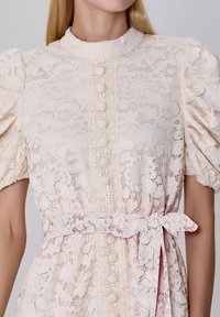 Robe en dentelle rose pâle avec des manches bouffantes, un col mandarin, une patte de boutonnage centrale et une ceinture en dentelle, ornée de motifs floraux.