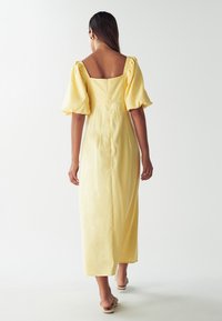 Žlté midi šaty s balónovými rukávmi, štvorcovým výstrihom na chrbte, textúrovanou tkaninou a skrytým zipsom, nosené s svetlými sandálmi.