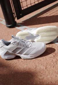 Witte Adidas tennisschoenen met grijze strepen liggen op een graveltennisbaan dichtbij een zwart netpaal en een witte baanlijn.