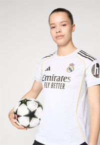 Maillot de football blanc avec des accents noirs et jaunes, comportant le logo "Emirates Fly Better" et le blason du Real Madrid. Un joueur tient un ballon de football noir et blanc à motifs.