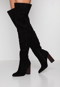 Botas de ante negras por encima de la rodilla con tacón cuadrado, que cuentan con una cremallera lateral y un diseño ligeramente holgado. Textura suave, puntera afilada.