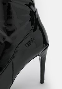 Botín de charol negro de tacón alto con un diseño elegante, que presenta una punta afilada y un logotipo en relieve en el lateral. Textura suave y brillante.