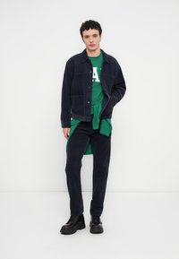 Σκούρο τζιν μπουφάν πάνω από πράσινο φούτερ με λευκά γράμματα, σε συνδυασμό με σκούρο παντελόνι και μαύρα μπότες με κορδόνια. Απλός σχεδιασμός, casual εμφάνιση.