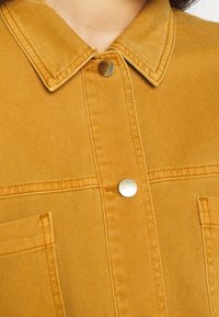 Žlutá džínová košile s límcem, dvěma předními kapsami a stříbrnými knoflíky. Materiál má jemný texturovaný vzor a viditelné švy.