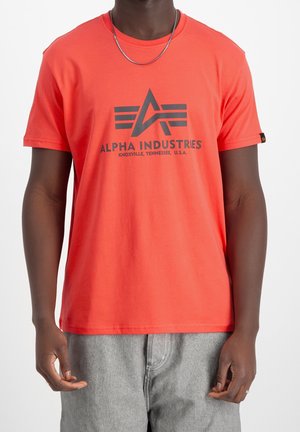 T-shirt en coton rouge avec un logo noir proéminent portant l'inscription « ALPHA INDUSTRIES » et un design à trois bandes en dessous, col rond, manches courtes.