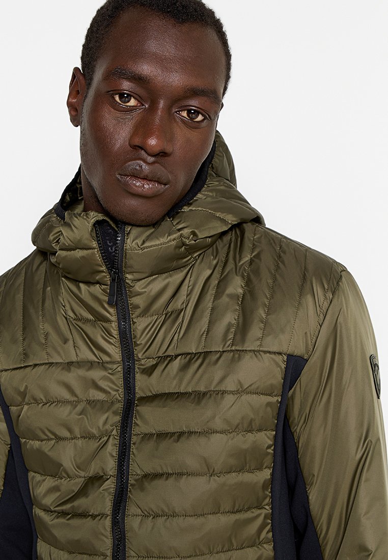 Olijfgroene gevoerde jas met capuchon, voorzien van een gequiltte textuur op het bovenlichaam en effen zwarte zijkanten, ritssluiting en een logo patch.
