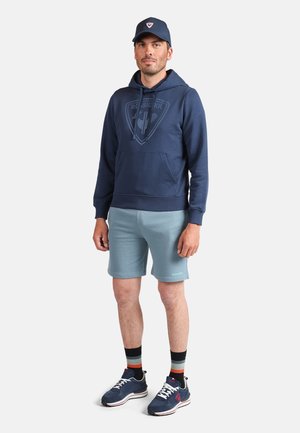 Marineblaues Hoodie mit Logo, hellblaue Shorts und marineblaue Sneaker mit roten Akzenten. Zubehör umfasst eine passende marineblaue Mütze und bunte Socken.