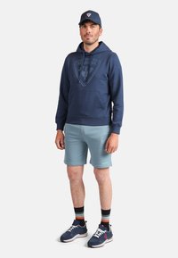 Námořnická mikina s kapucí a logem, světle modré kraťasy, černo-červené tenisky a modrá čepice. Mikina má šňůrky a přední kapsu.