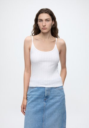 Jeune femme aux longs cheveux bouclés bruns, portant un haut blanc sans manches et une jupe en jean bleu clair, debout devant un fond uni.