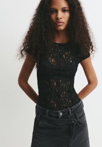 Junge Frau mit langen, lockigen Haaren, die ein enges schwarzes Spitzenoberteil und einen hoch taillierten schwarzen Jeansrock vor einem schlichten Hintergrund trägt.