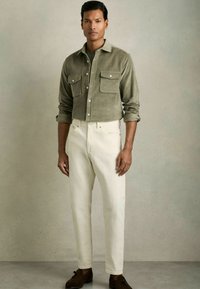 Cămașă verde din corduroy cu două buzunare pe piept, asortată cu pantaloni albi cu talie înaltă și pantofi din suede maro. Fundal neutru.
