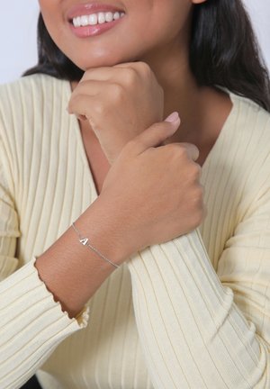 Bracciale in argento con un charm a forma di "A", indossato su un polso abbronzato. Chi lo indossa ha un maglione a coste di un leggero colore giallo.