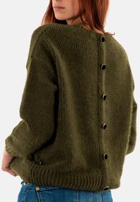 Oliven grøn strikket sweater med løs pasform, der har en høj krave og en række sorte knapper langs ryggen. Struktureret garn og ribbet kant.