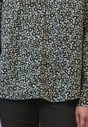 Koszula zapinana na guziki z gęstym wzorem małych czarnych i żółtych kwiatów na stonowanym niebiesko-zielonym tle, zestawiona z czarnymi spodniami.