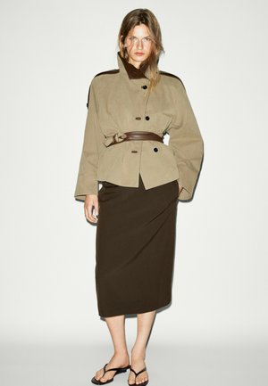 Vrouw draagt een beige jasje met riem, een donkerbruine midirok en zwarte slippers, staand tegen een effen witte achtergrond.