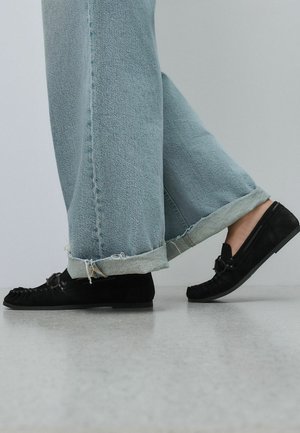 Henkilö kävelee vaaleansinisissä farkuissa, joissa on rispaantuneet reunat, ja mustissa mokkasiinityylisissä loafer-kenkäparissa sileällä harmaalla lattialla.