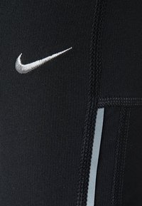 Czarny elastyczny materiał z czarnym przeszyciem i małym białym logo Nike w kształcie skrzydła nad pionowym odblaskowym paskiem.