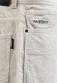 White corduroy fabric pants featuring vertical ribbing, with five pockets and embroidered "WASTED" logo in dark thread.