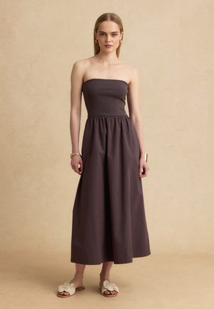 Femme portant une robe longue marron foncé sans bretelles avec une taille froncée, des boucles d'oreilles créoles dorées, des bracelets et des sandales plates beiges texturées.