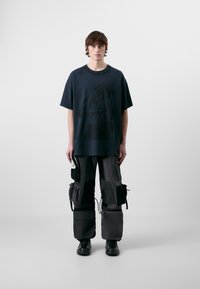 Camiseta gráfica oscura de corte holgado, combinada con pantalones cargo que cuentan con múltiples bolsillos y correas ajustables. Botas negras completan el look.