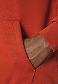 Red hoodie featuring a front pocket, ribbed cuffs, and textured fabric. A hand is resting inside the pocket, showcasing the material's stretch.