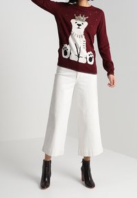 Maglione bordeaux con design di un orso polare bianco e corona, abbinato a pantaloni bianchi ampi e stivaletti neri alla caviglia. Materiale in maglia testurizzata.
