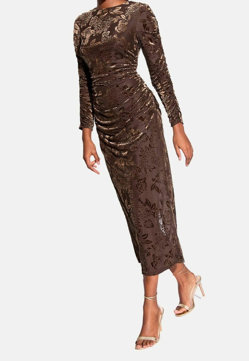 Femme portant une robe midi marron à manches longues, à motif fleuri et froncée, et des sandales à talons hauts beiges, debout devant un fond uni.