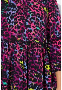 Schwarzes Oberteil aus Stoff mit einem bunten Leopardenmuster in Pink, Blau und Grün. Verfügt über einen gerafften Bund und kurze Ärmel.