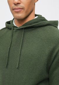 Olive zöld kapucnis pulóver, texturált anyagból készült, kenguruzsebbel, húzózsinórokkal és rávarrt ujjal, a laza illeszkedés érdekében.