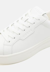 Hvit sneaker med en glatt syntetisk overdel, rund tå, flate snørebånd, preget merkevarelogo og en tykk gummisåle med subtile riller.