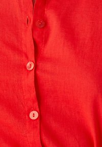 Camisa de lino roja con una textura suave, cierre de botones y sutiles detalles de costura a lo largo de la tapeta.