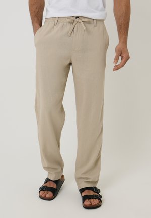 Mand iført beige bukser med snor og sorte sandaler med spænde, stående med afslappede hænder mod en ensartet lys baggrund.