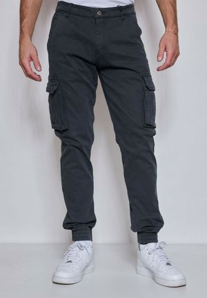 Homme portant un pantalon cargo jogging sombre et des baskets blanches, debout contre un mur uni avec les mains détendues le long du corps.