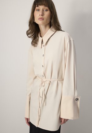 Femme aux cheveux bruns portant une chemise beige clair à manches longues, boutonnée, avec de larges poignets et une ceinture nouée à la taille, devant un fond uni.