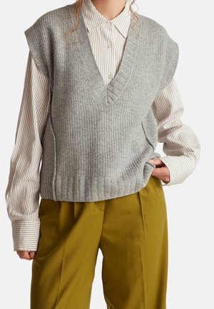 Personne portant un pull sans manches en tricot gris à col en V par-dessus une chemise blanche et marron rayée à boutons avec un pantalon jaune moutarde.