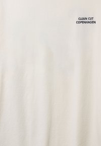 Valkoinen puuvillainen t-paita, jossa on lyhyet hihat ja pyöreä pääntie. Edessä on tummansinisin kirjaimin painettu teksti "CLEAN CUT COPENHAGEN".