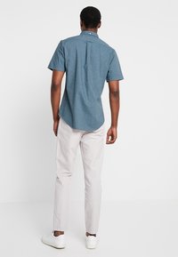 Chemise à manches courtes couleur sarcelle avec col boutonné et pli dans le dos, associée à un pantalon clair et des baskets blanches.