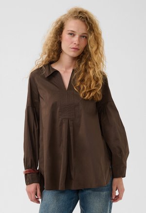 Vrouw met krullend blond haar, een losse donkerbruine blouse met lange mouwen en blauwe spijkerbroek tegen een effen achtergrond.