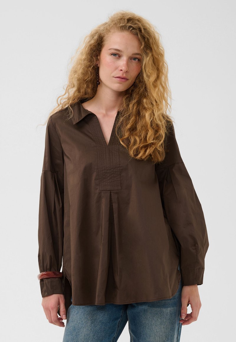 Femme aux cheveux blonds bouclés portant un chemisier ample à manches longues marron foncé et un jean bleu sur un fond uni.