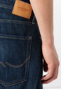 Jeans de mezclilla oscura con una etiqueta de cuero que dice "JACK & JONES" en la parte trasera. Presentan costuras amarillas y un diseño de pierna ligeramente ajustada.