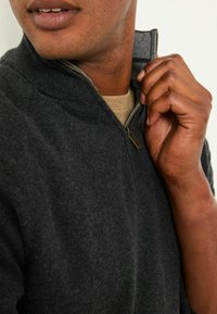 Dunkelgrauer Reißverschlusspullover mit strukturierter Strickware, ausgestattet mit einem hohen Kragen und goldenen Reißverschlussdetails. Der innere Kragen zeigt einen kontrastierenden Stoff.