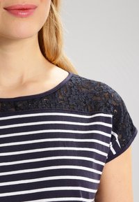 Navy ja valkoisia raitoja oleva paita, jossa on pitsikoristelu harteilla. Lyhyet hihat ja pyöreä kaula-aukko. Pehmeä kankaan rakenne.