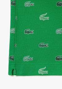 Tissu vert avec de petits logos crocodile brodés vert foncé répétitifs, dispersés sur la surface près d'un bord cousu avec une petite fente latérale.