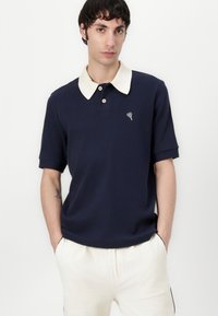 Námornícke polo tričko s krémovým golierom, krátkymi rukávmi a malým srdiečkovým logom. Textúrovaná látka s gombíkovým zapínaním a voľným strihom.